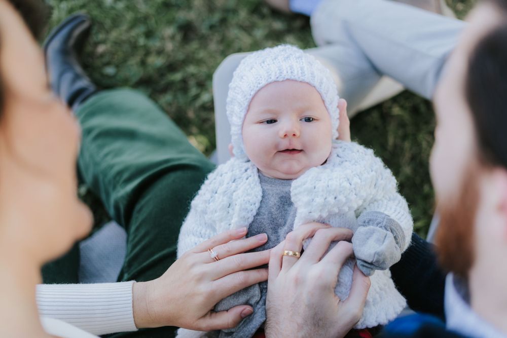 Baby in white knit outfit held by parents outdoors.