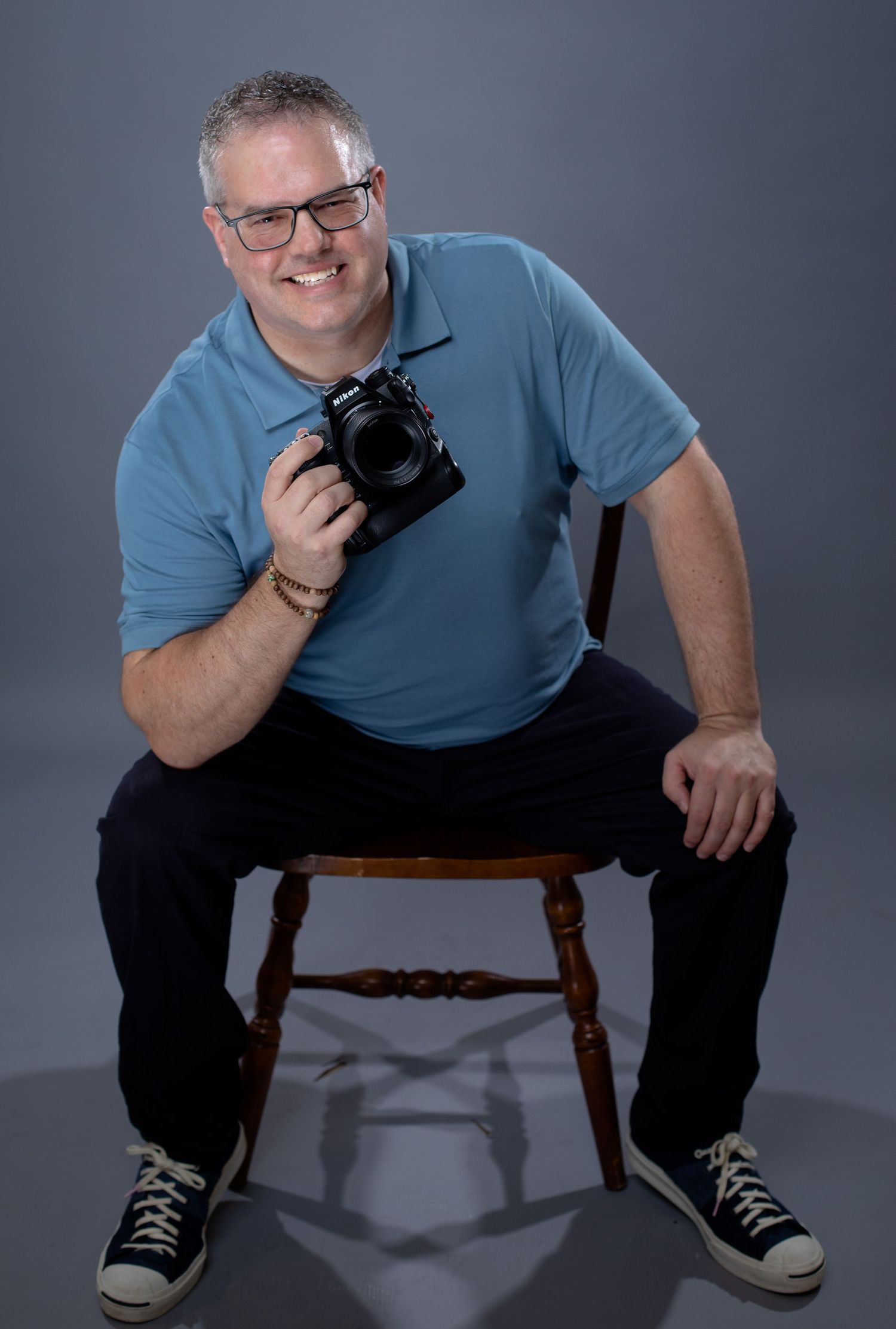 Portrait of photographer in blue polo shirt sitting on wooden stool with camera.