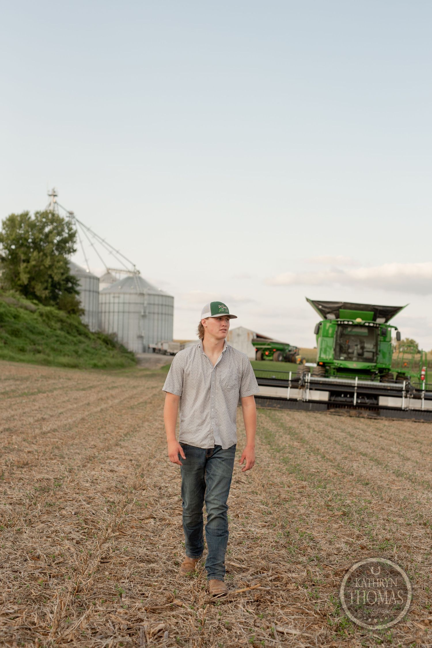 Person walking through harvested field with grain silos and farm equipment in background.
