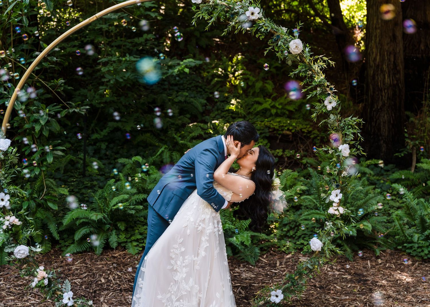 A romantic dip kiss captured under forest trees with sunlight filtering through.