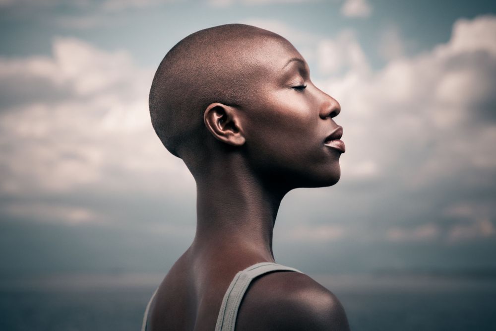 Portrait of a bald woman with shut eyes amid a cloudy marine landscape, shot in an ad-style photoshoot.