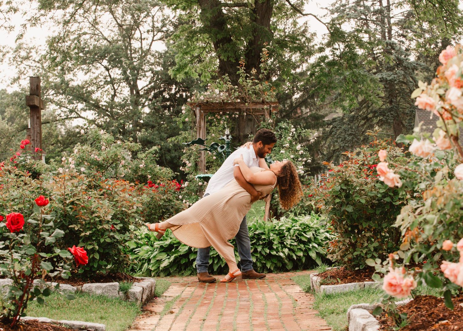 A couple shares a romantic dip kiss on a garden path surrounded by colorful blooming roses.