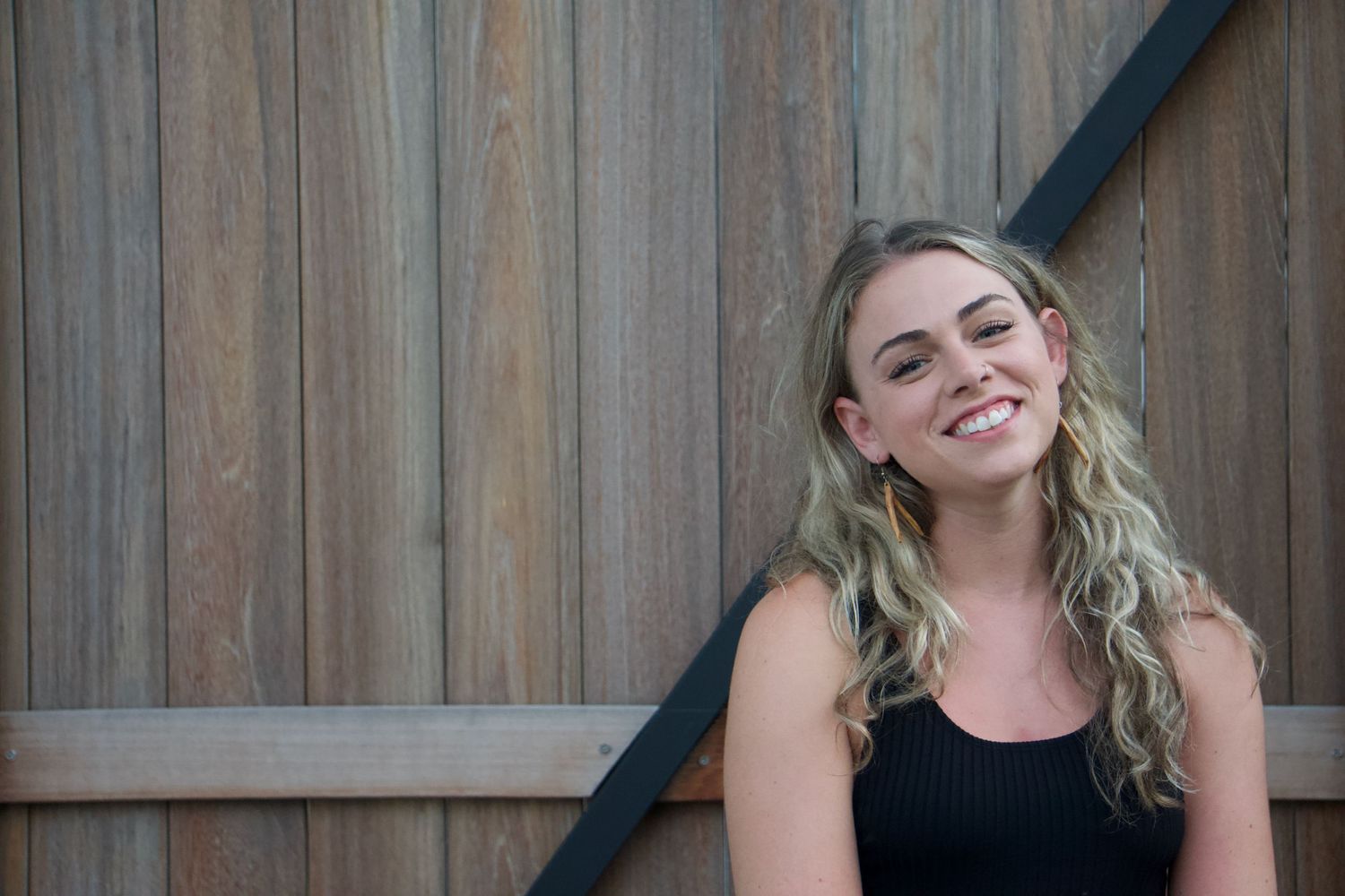 A woman in a black shirt in front of a wood and iron gate smiles with blonde curly hair and handmade clay earrings