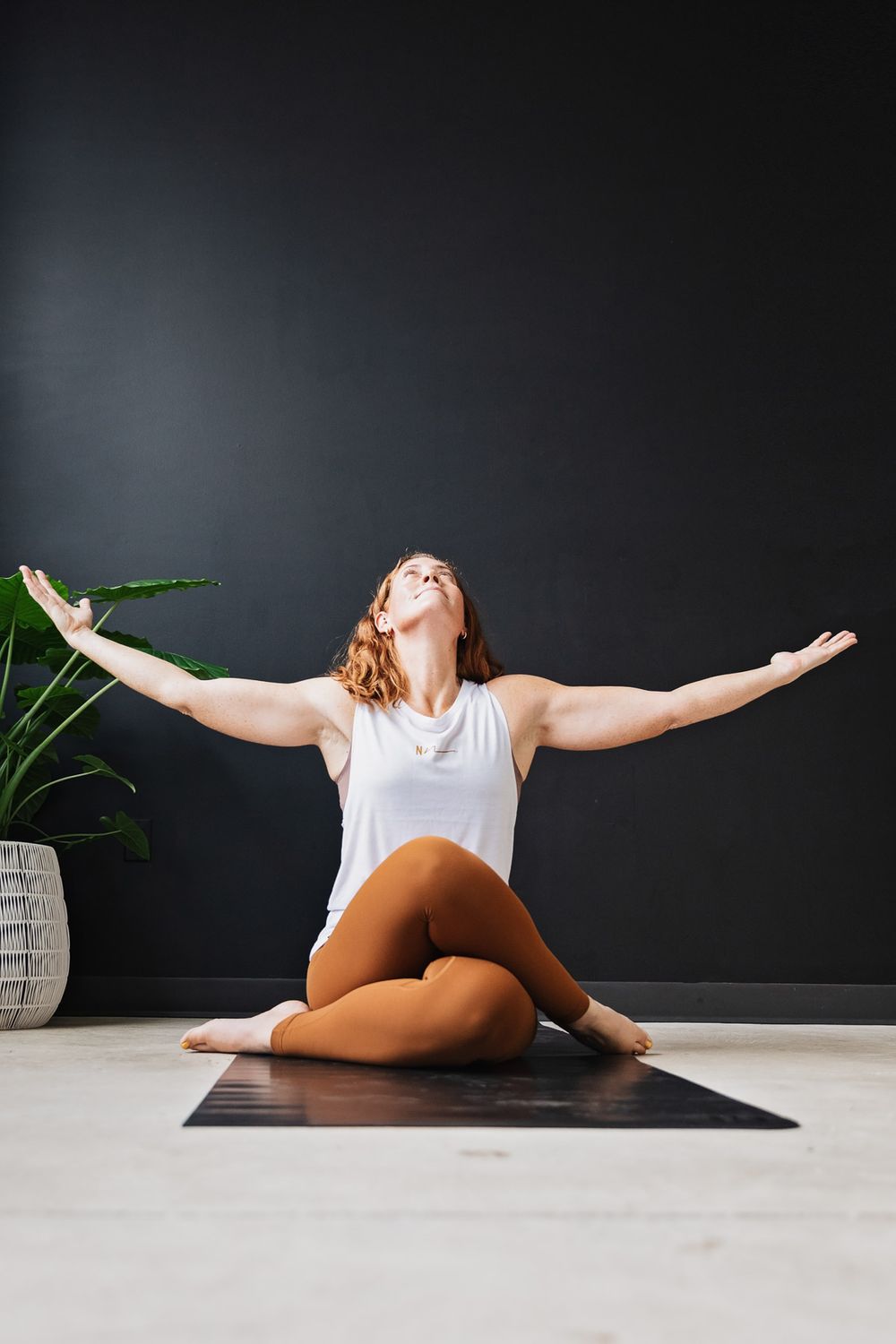Expressive yoga pose with arms outstretched on black mat against dark wall.