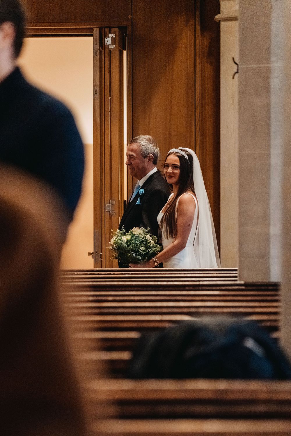 A bride and her escort walk down a wooden church aisle carrying a blue floral bouquet.
