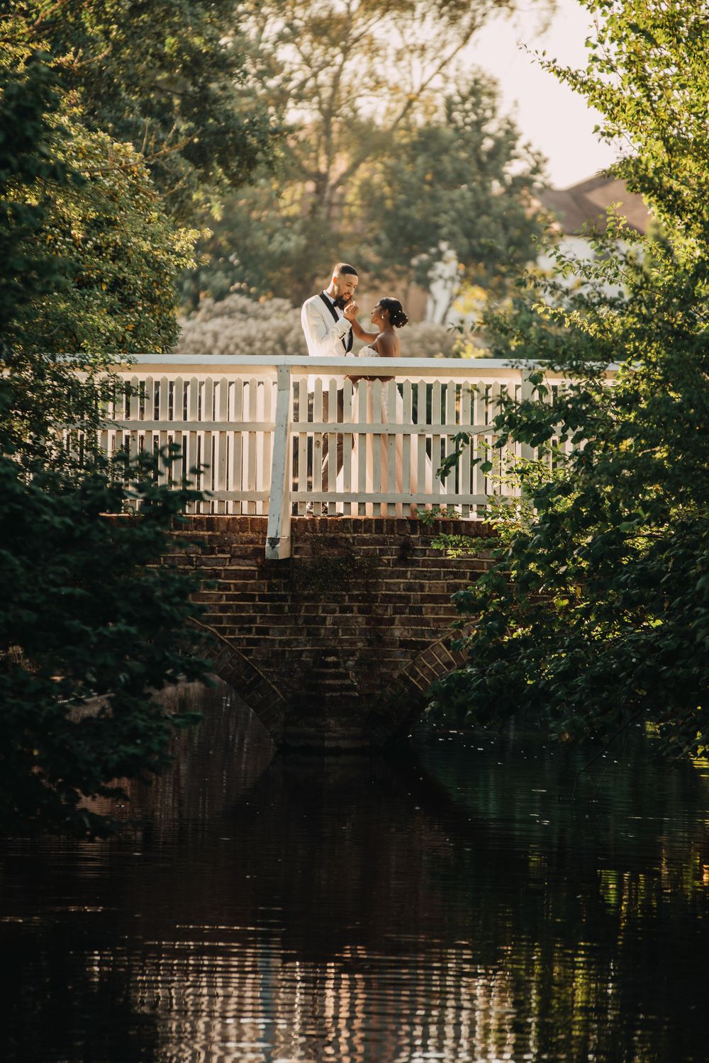 Newlyweds share an intimate moment on a white bridge over a reflective pond at sunset.