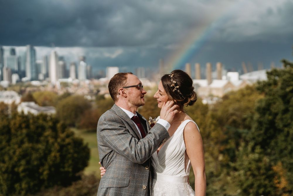 A wedding couple embraces with a dramatic cityscape and rainbow in the background.