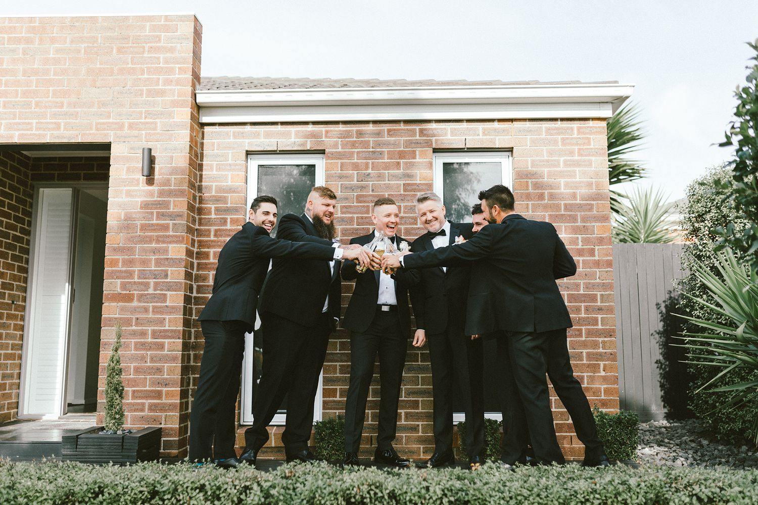 Wedding party in formal attire poses casually against brick wall of building.