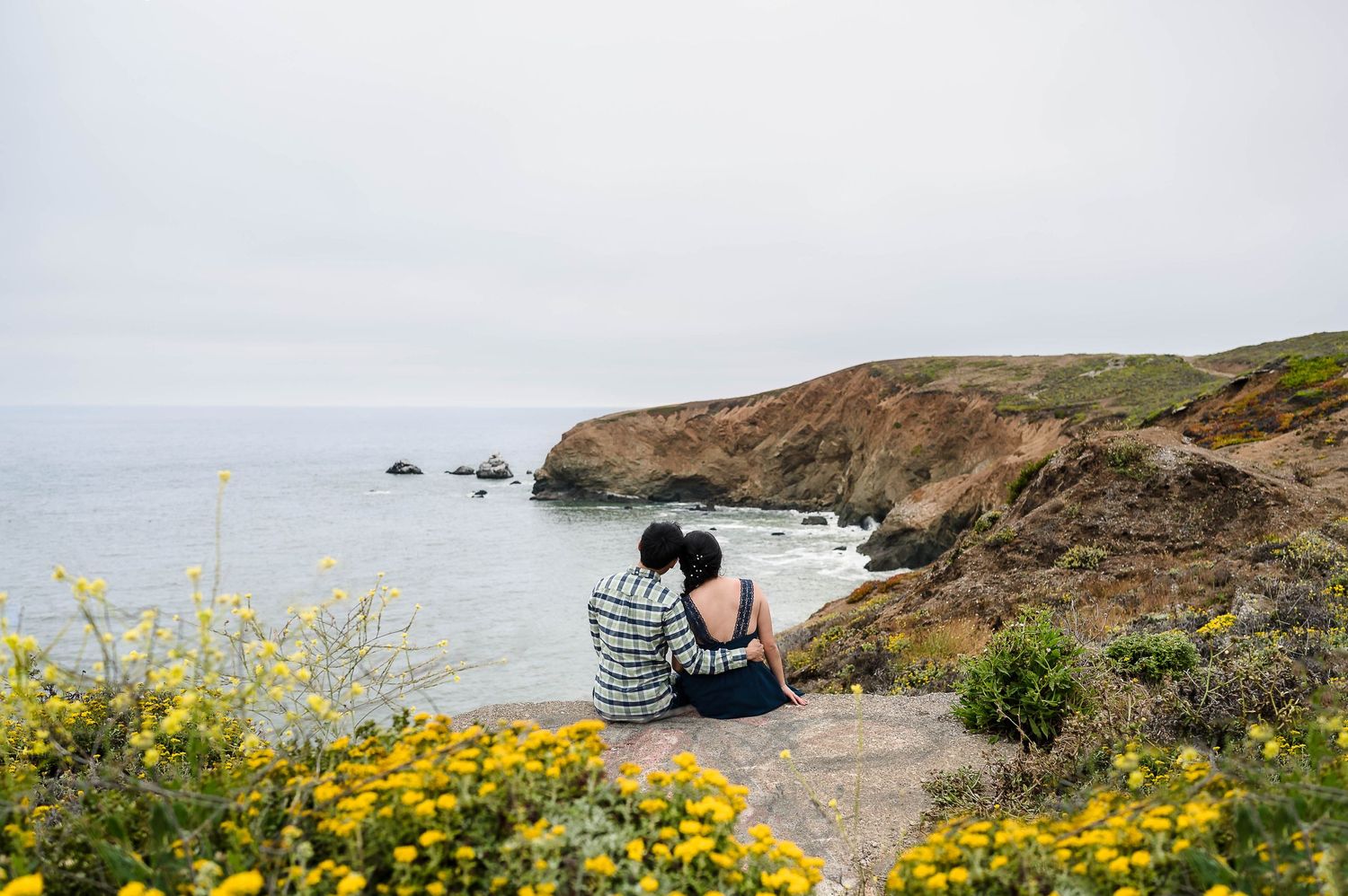 Two people sitting on a cliff overlooking the ocean with yellow wildflowers in the foreground.
