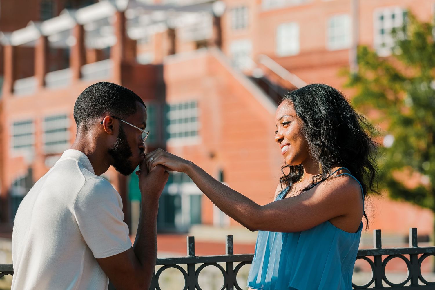 A man kisses a smiling woman's hand outdoors near an iron fence with a brick building behind them.