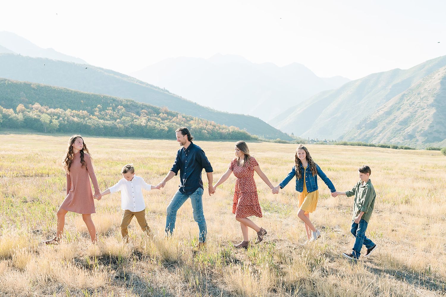 Six people join hands while walking through a sunny mountain valley with peaks in the background.