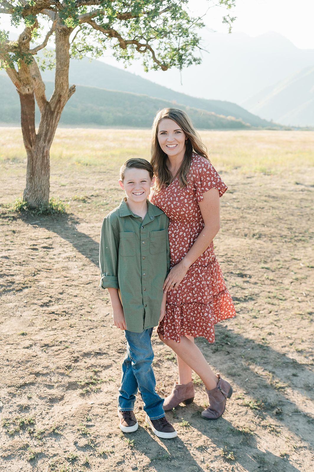 A casual outdoor portrait beneath a tree with mountains in the background on a sunny day.