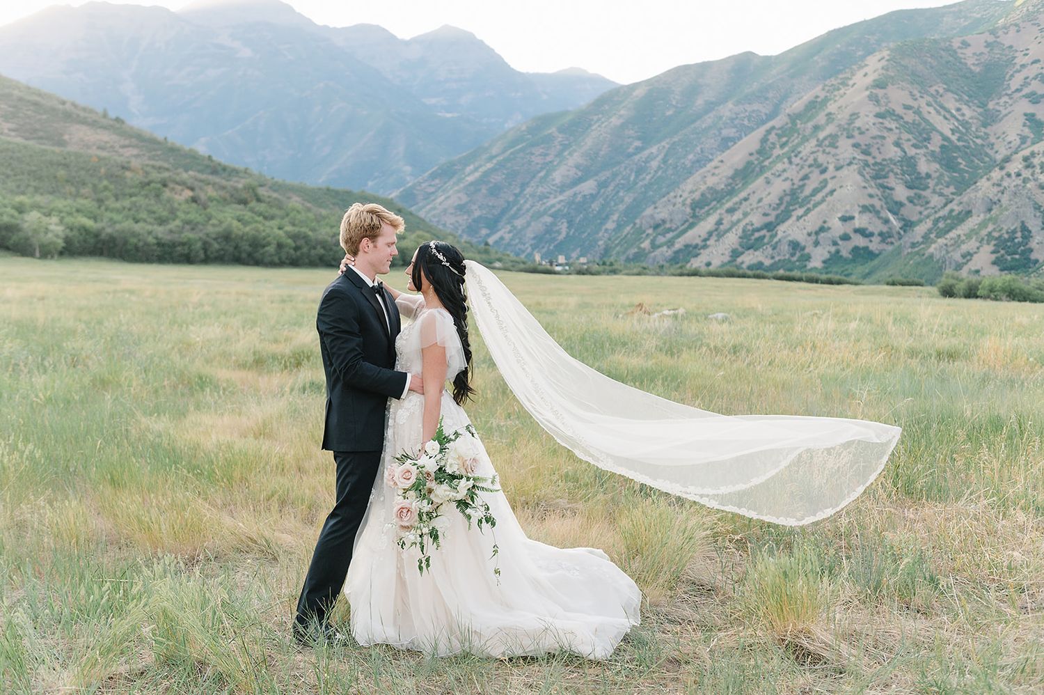 Wedding couple embracing in a mountain meadow with a flowing veil catching the breeze.