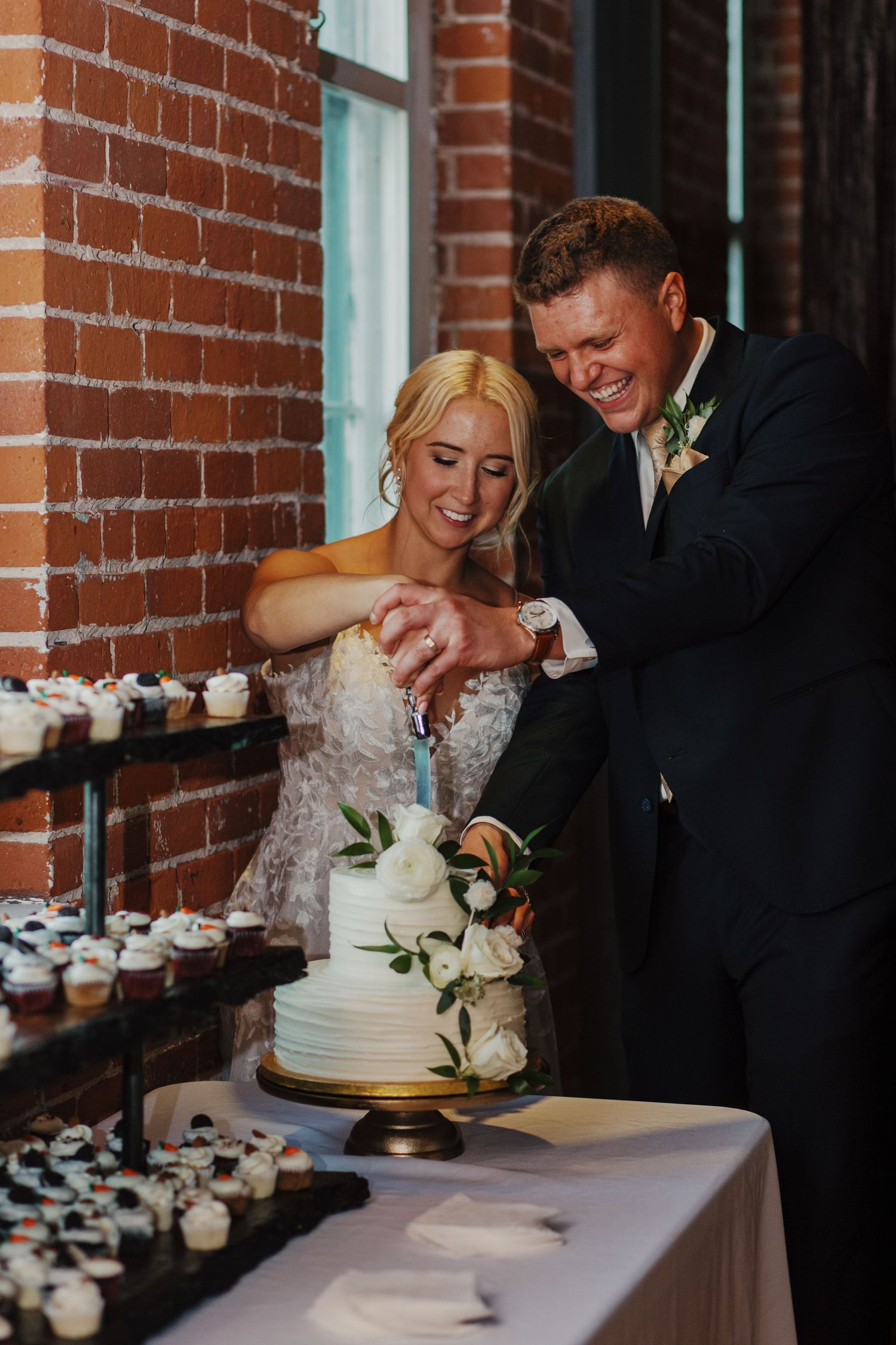 Newlyweds cutting their elegant white wedding cake with cupcakes displayed.