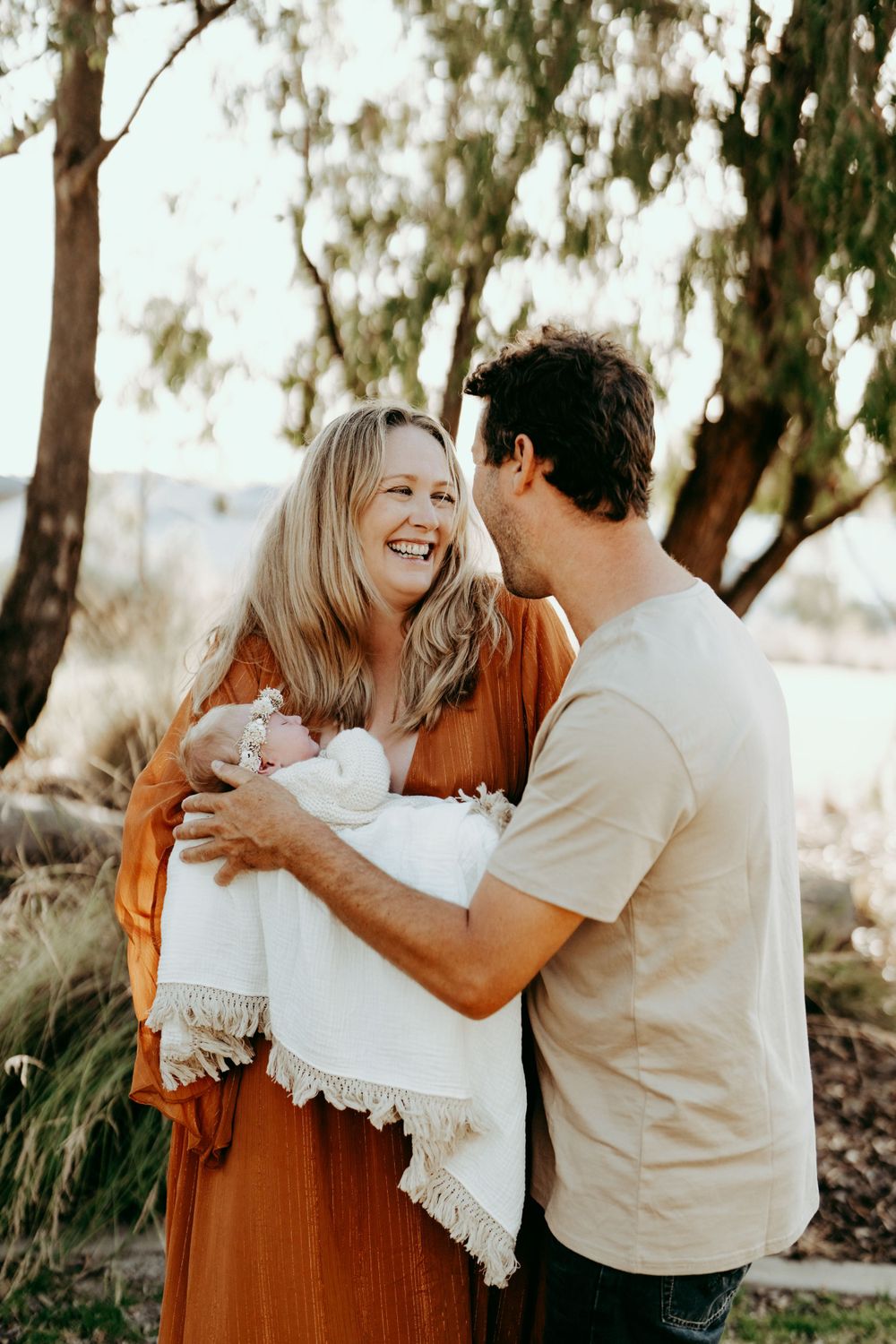 Couple sharing joyful moment while holding baby wrapped in white blanket outdoors.