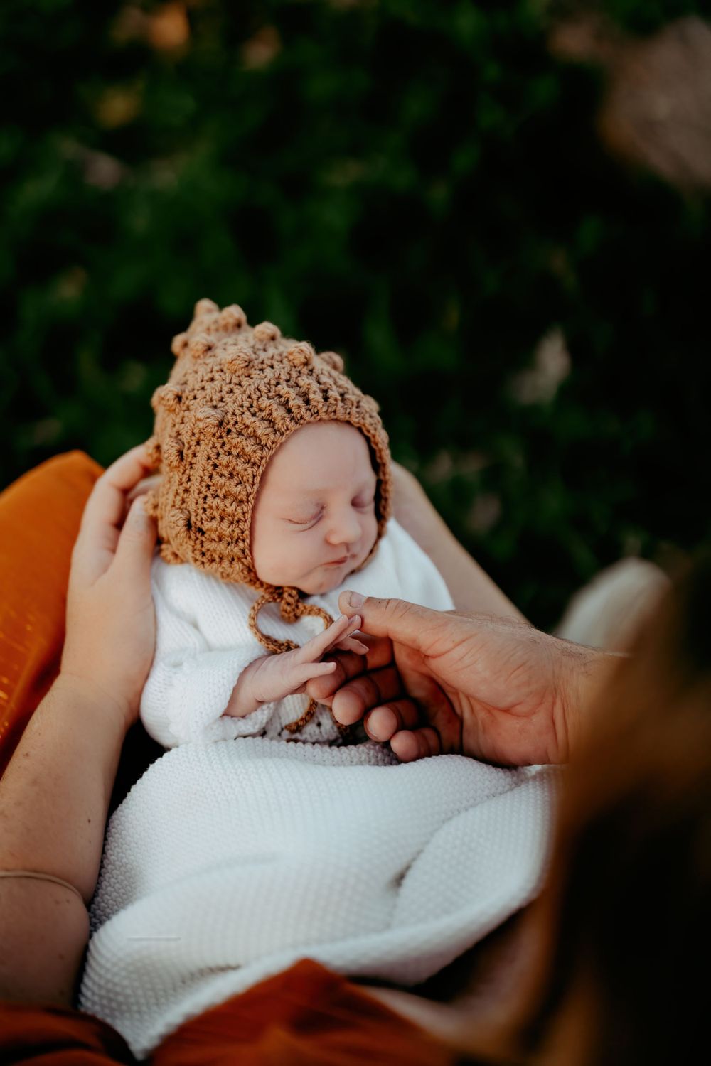 Sleeping newborn wearing knit bonnet swaddled in white blanket outdoors.