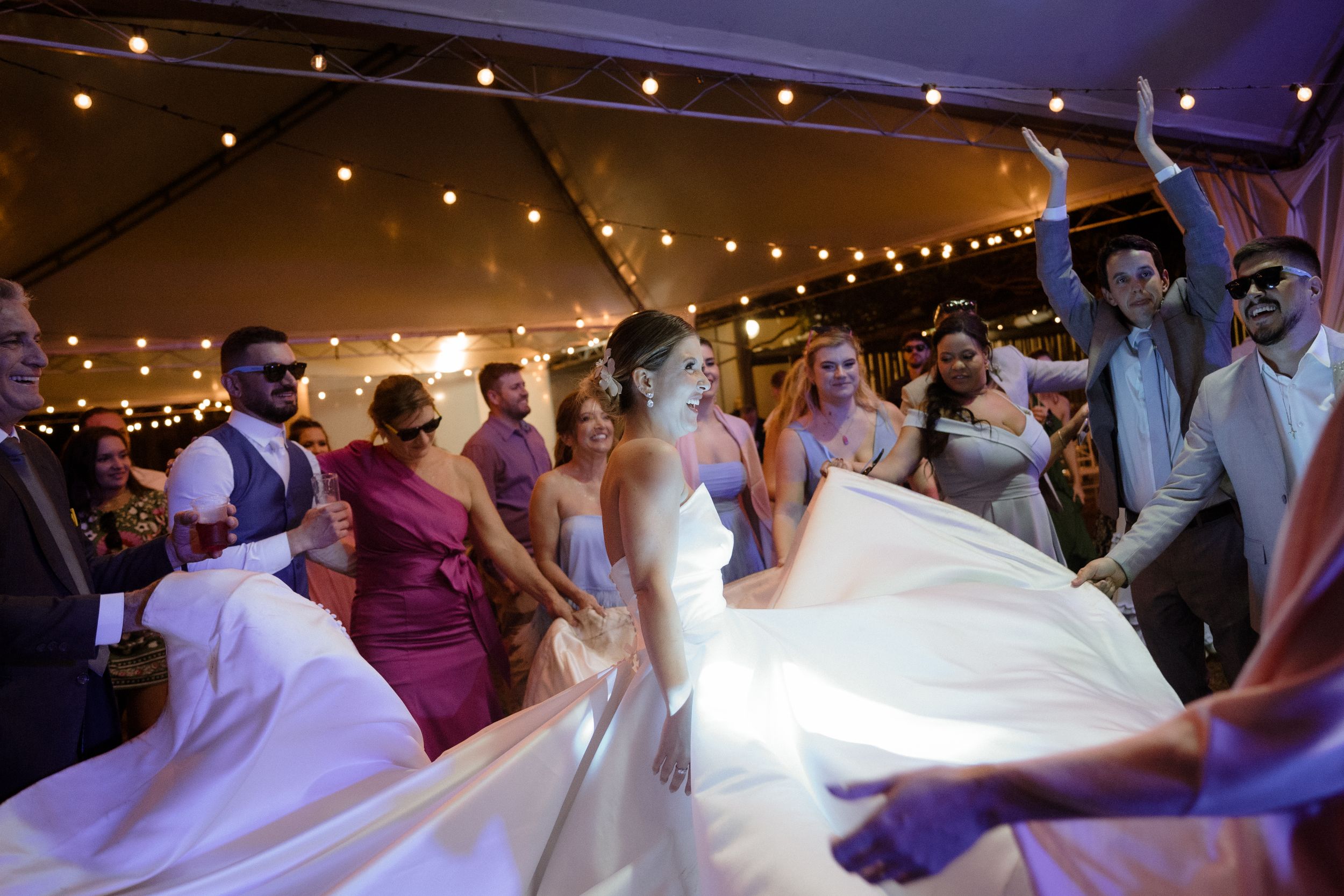 Wedding party dancing under tent with string lights and flowing white dress.