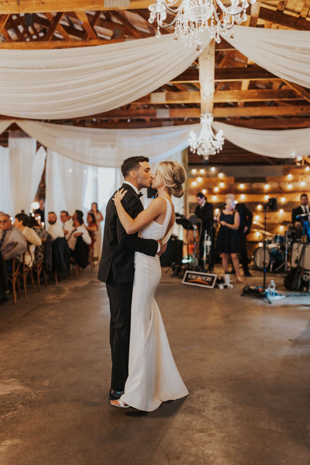 First dance at wedding reception in rustic barn venue with draped white fabric and chandeliers.