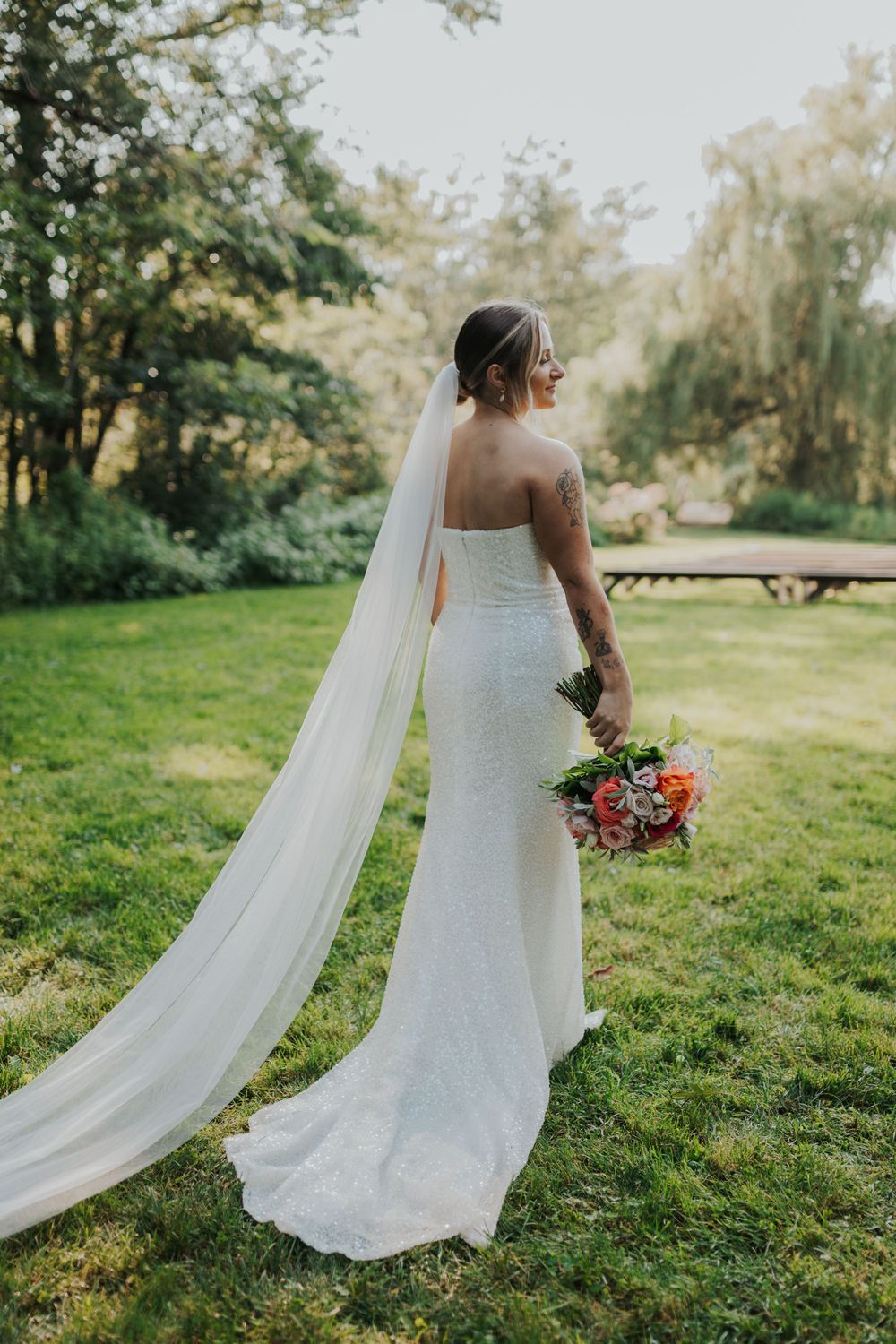 Bride in fitted wedding dress with long veil holds coral bouquet in garden setting.
