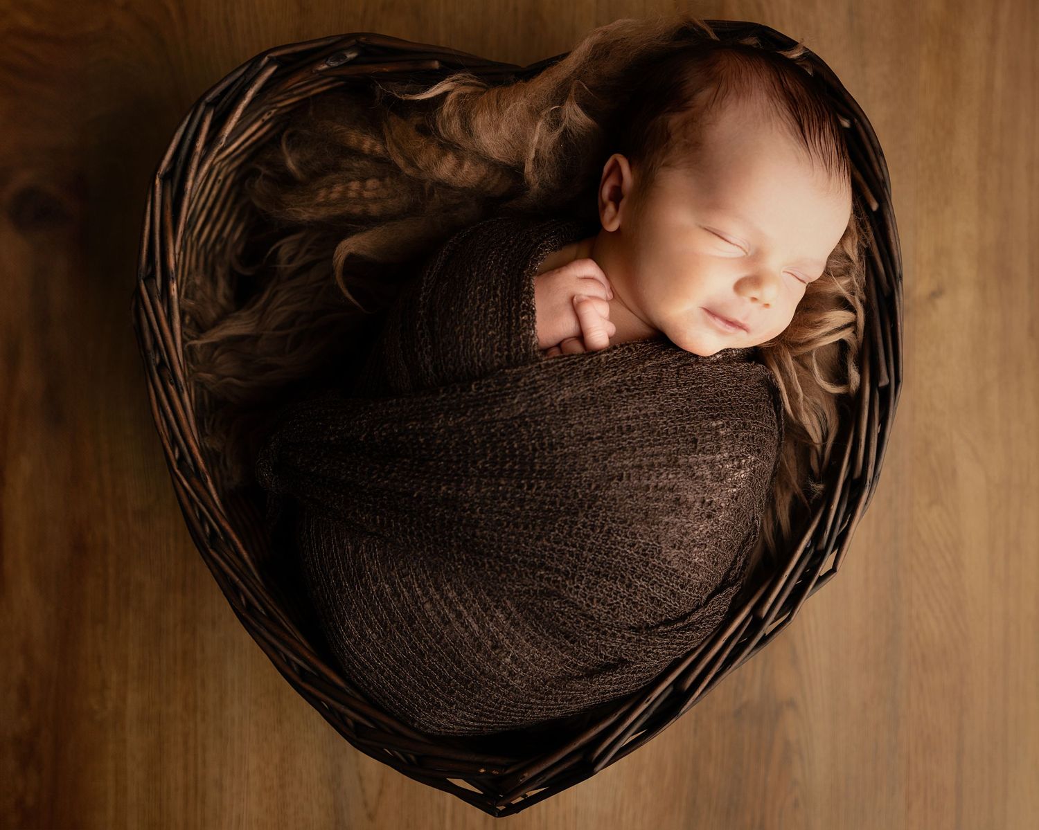 Newborn baby sleeping peacefully wrapped in brown swaddle blanket inside heart-shaped wicker basket.