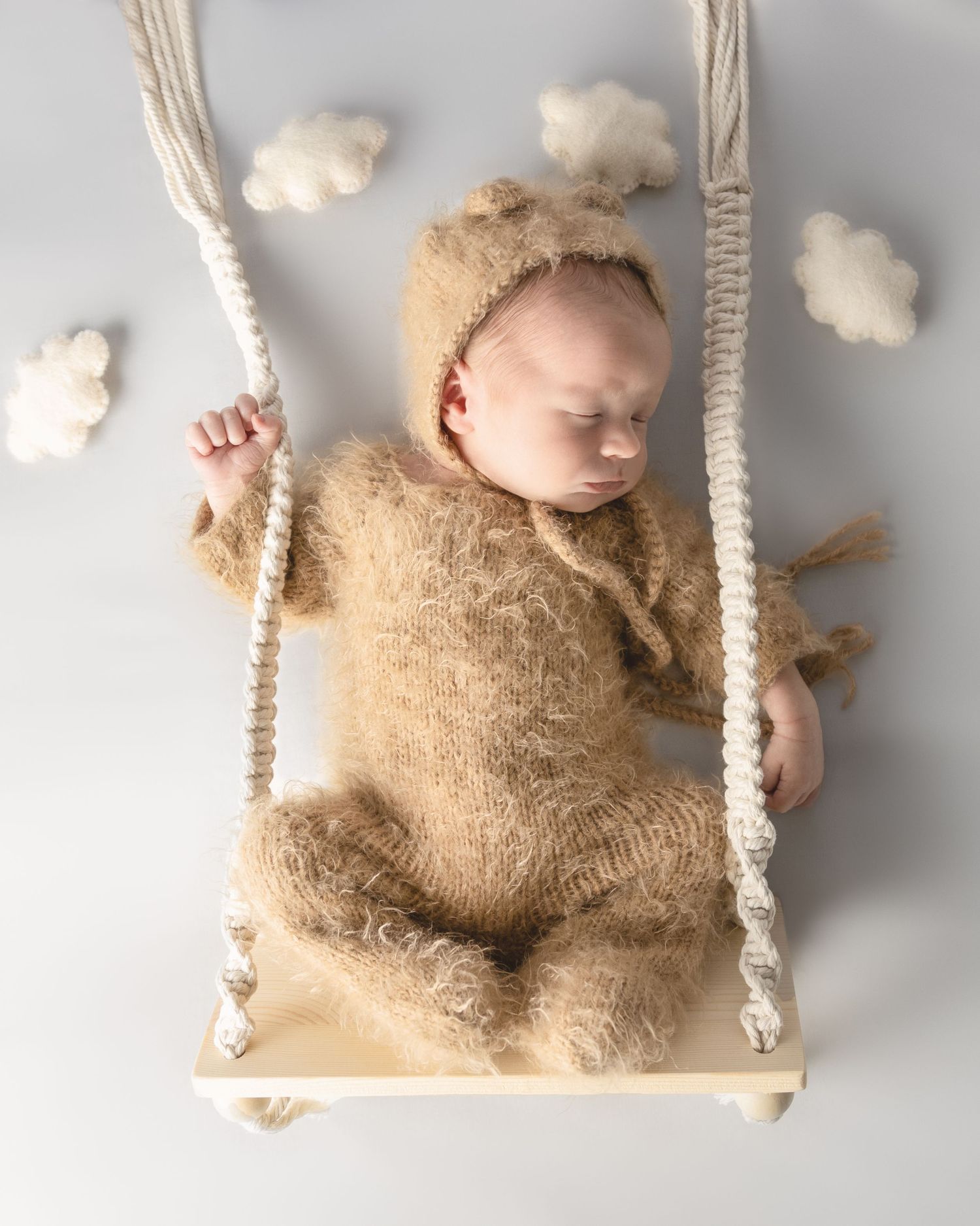Sweet newborn photography with baby in cozy beige knit outfit sleeping on tiny wooden swing with cotton cloud decor.
