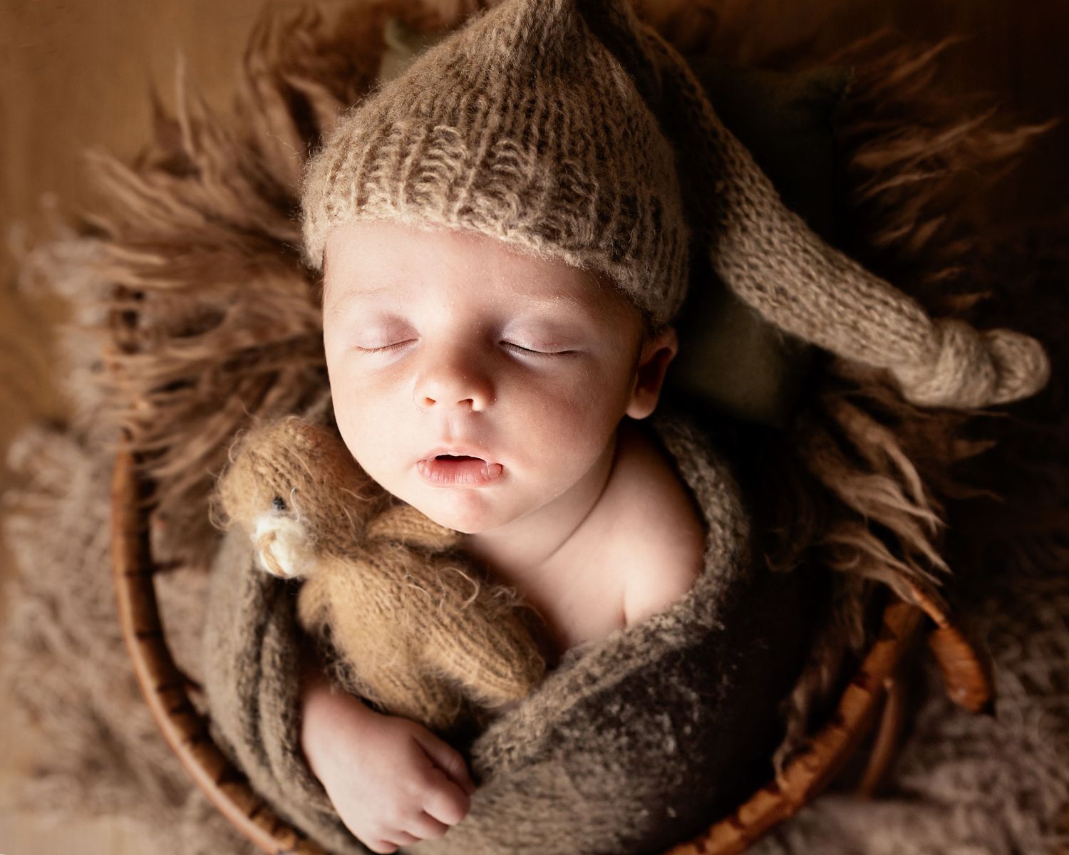 A sleeping baby wearing a knit hat lies peacefully wrapped in brown textured blankets on a soft furry backdrop.