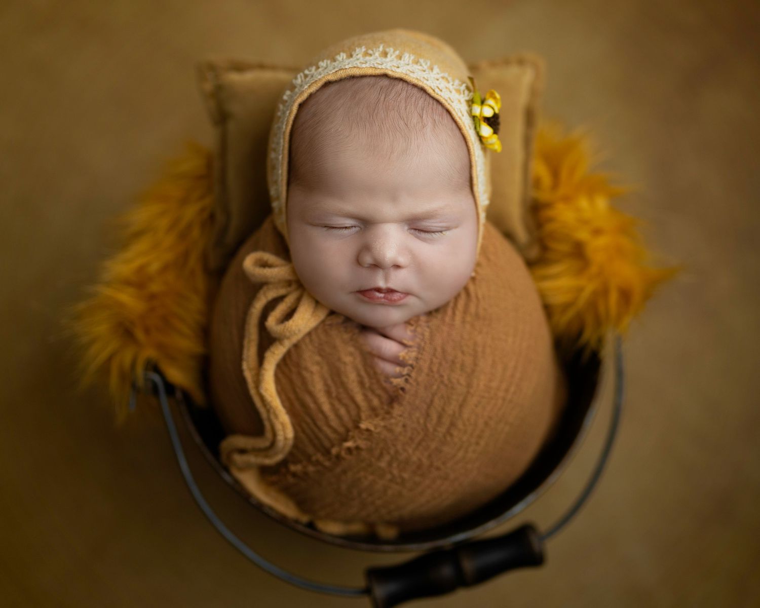 Newborn baby peacefully sleeping in mustard swaddle wrap with lace bonnet and yellow flower in circular wooden prop.