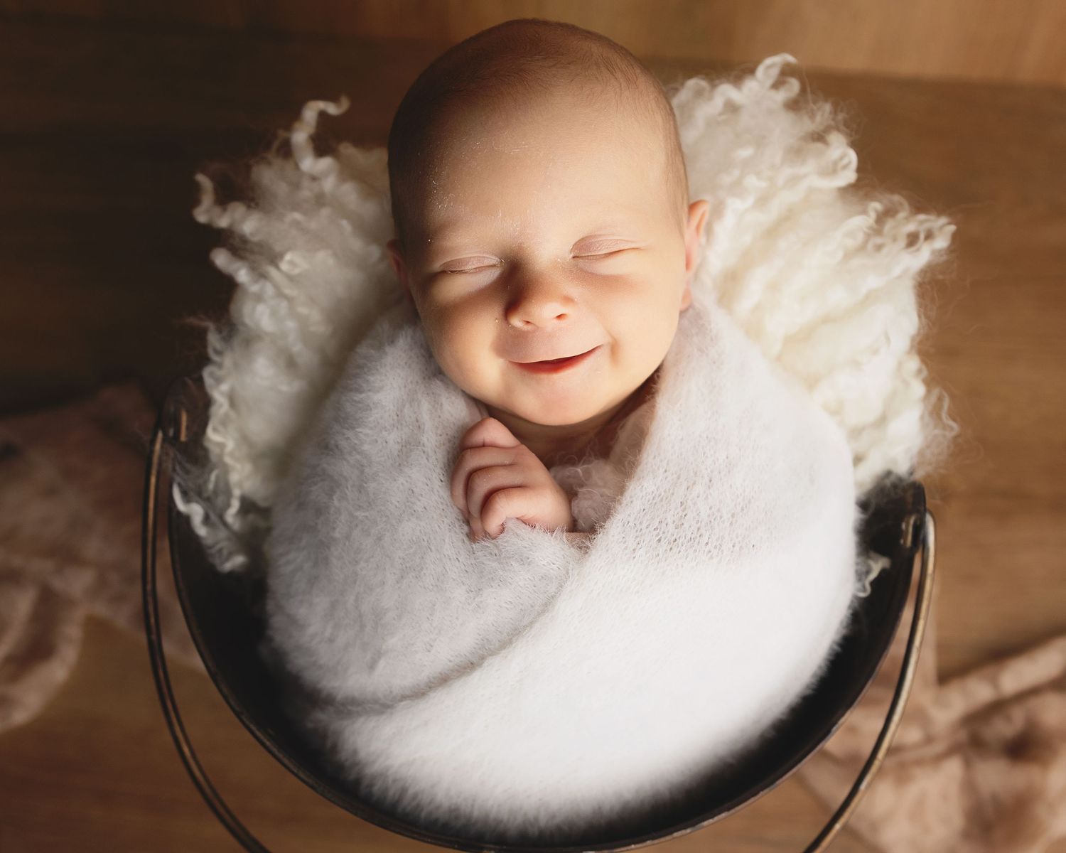 Sleeping newborn wrapped in white cloth lying in a dark metal bucket with soft lighting surrounding them.