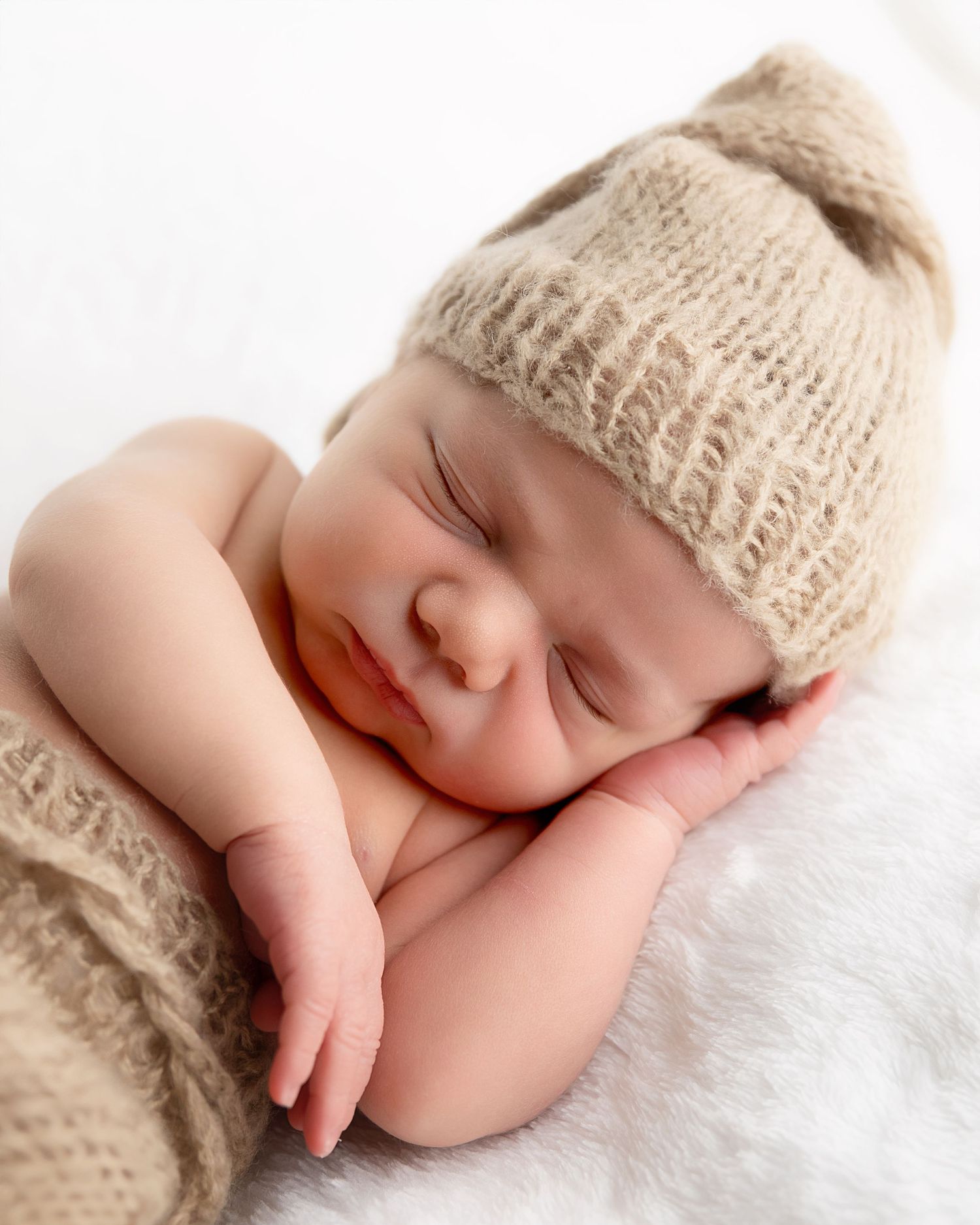 Peacefully sleeping baby wearing a cozy knit beanie and wrap on soft white blanket.