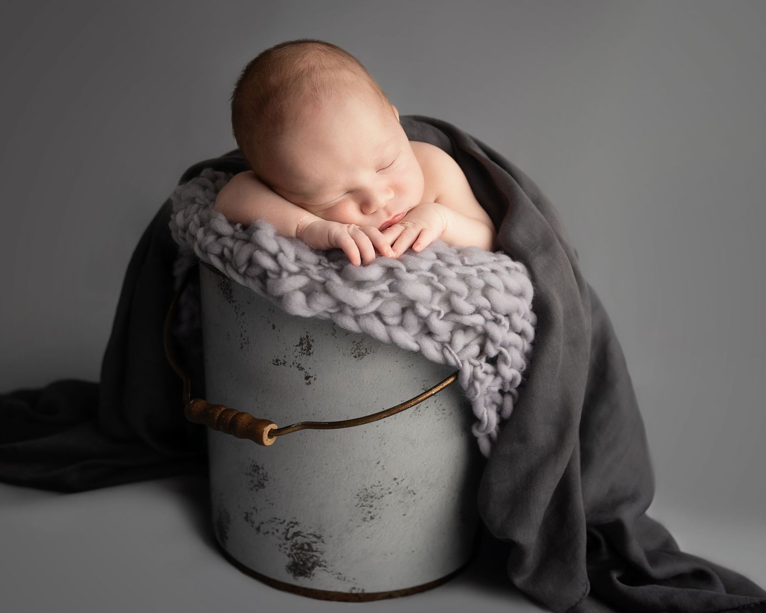Sleeping newborn rests on a grey fluffy blanket inside a rustic metal bucket in a studio setting.