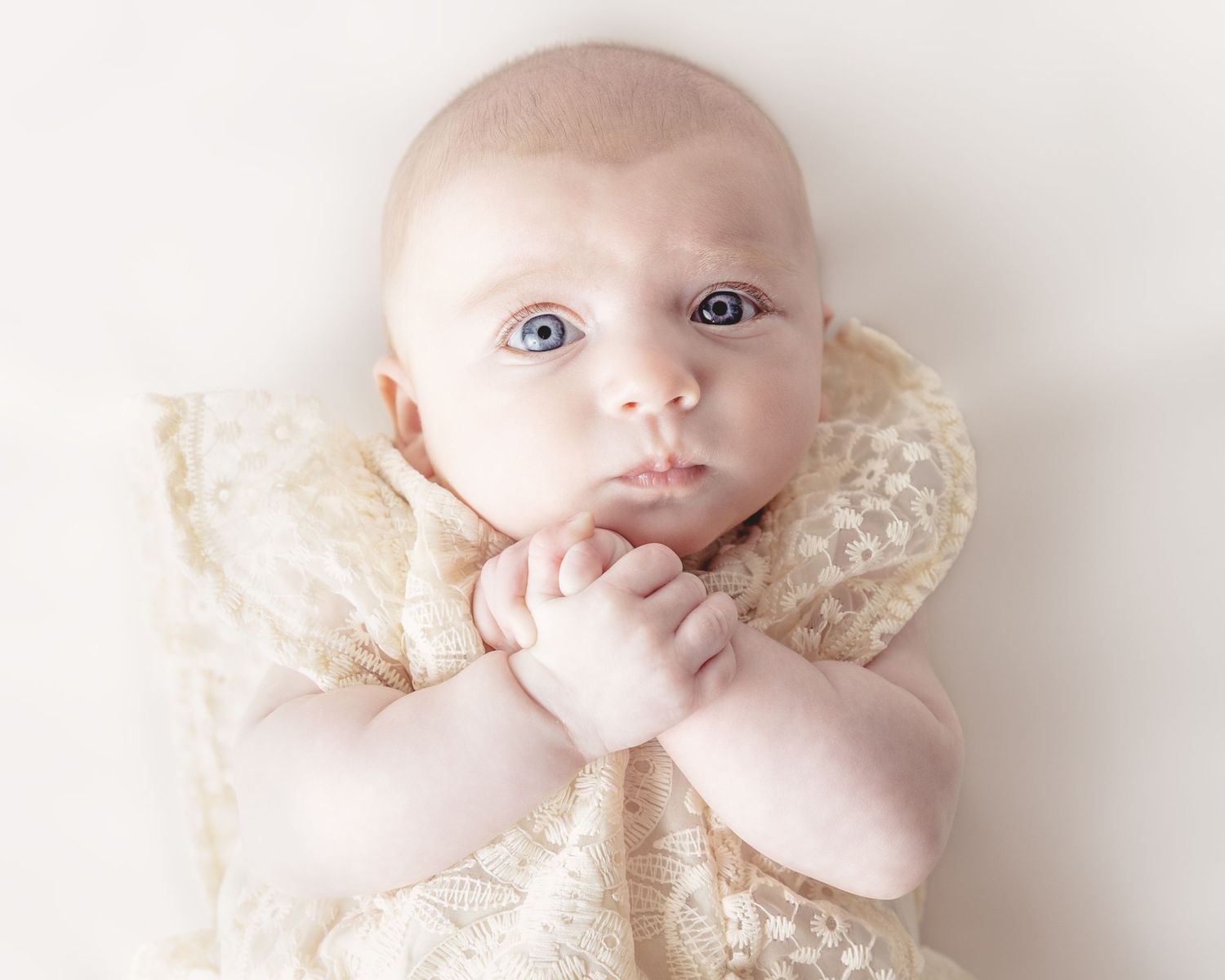 A cherubic baby in a vintage lace dress lies gazing upward with hands clasped under chin.
