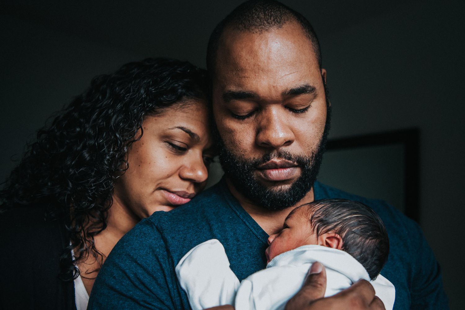 A woman leans her head on her husbands shoulder as they both look at their newborn in his arms.
