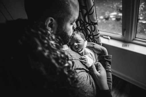 A man holds his newborn son in his arms in front of a window as his wife wraps her arms around him.
