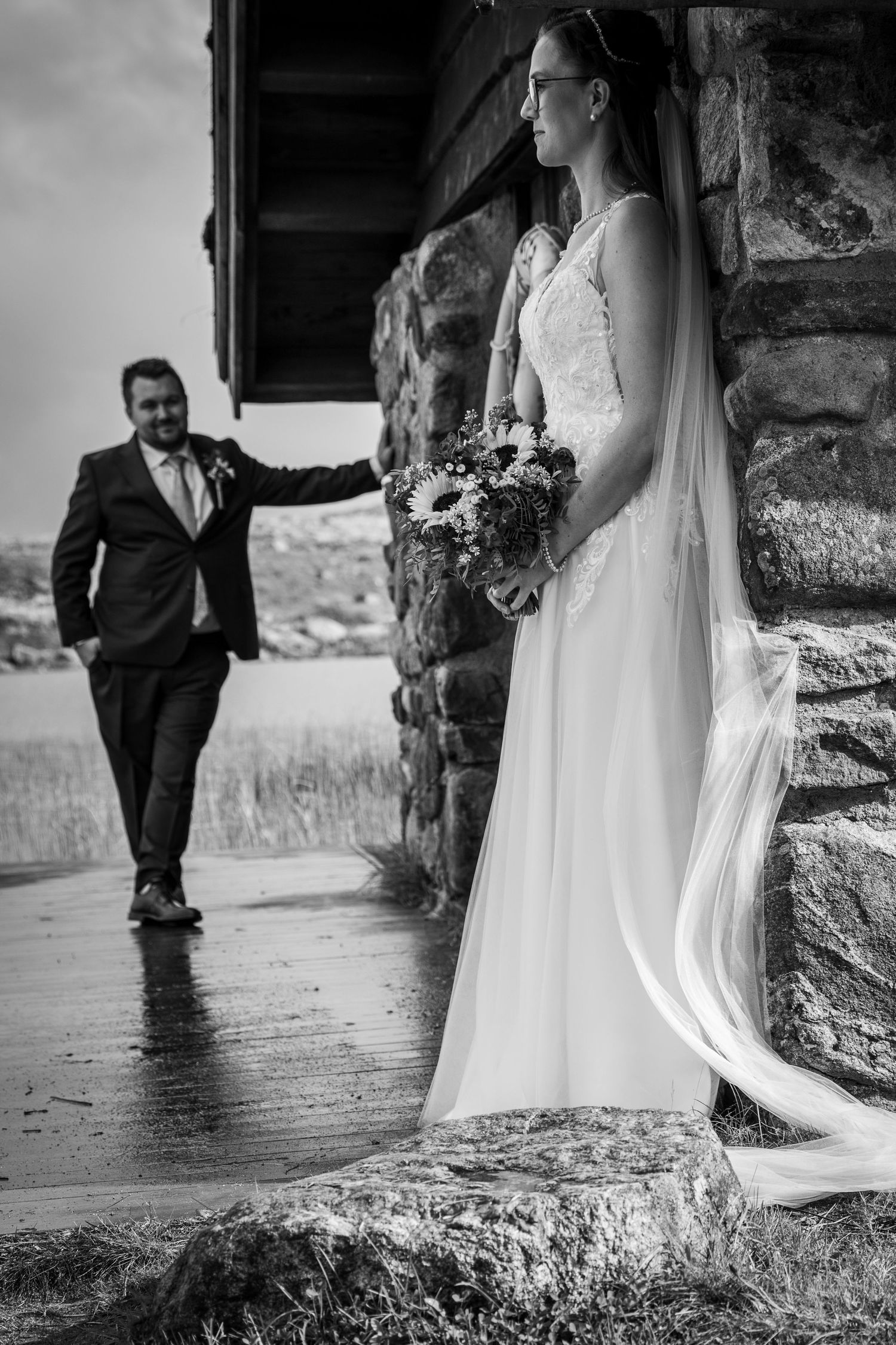 Black and white photo of bride with bouquet and groom approaching stone building.