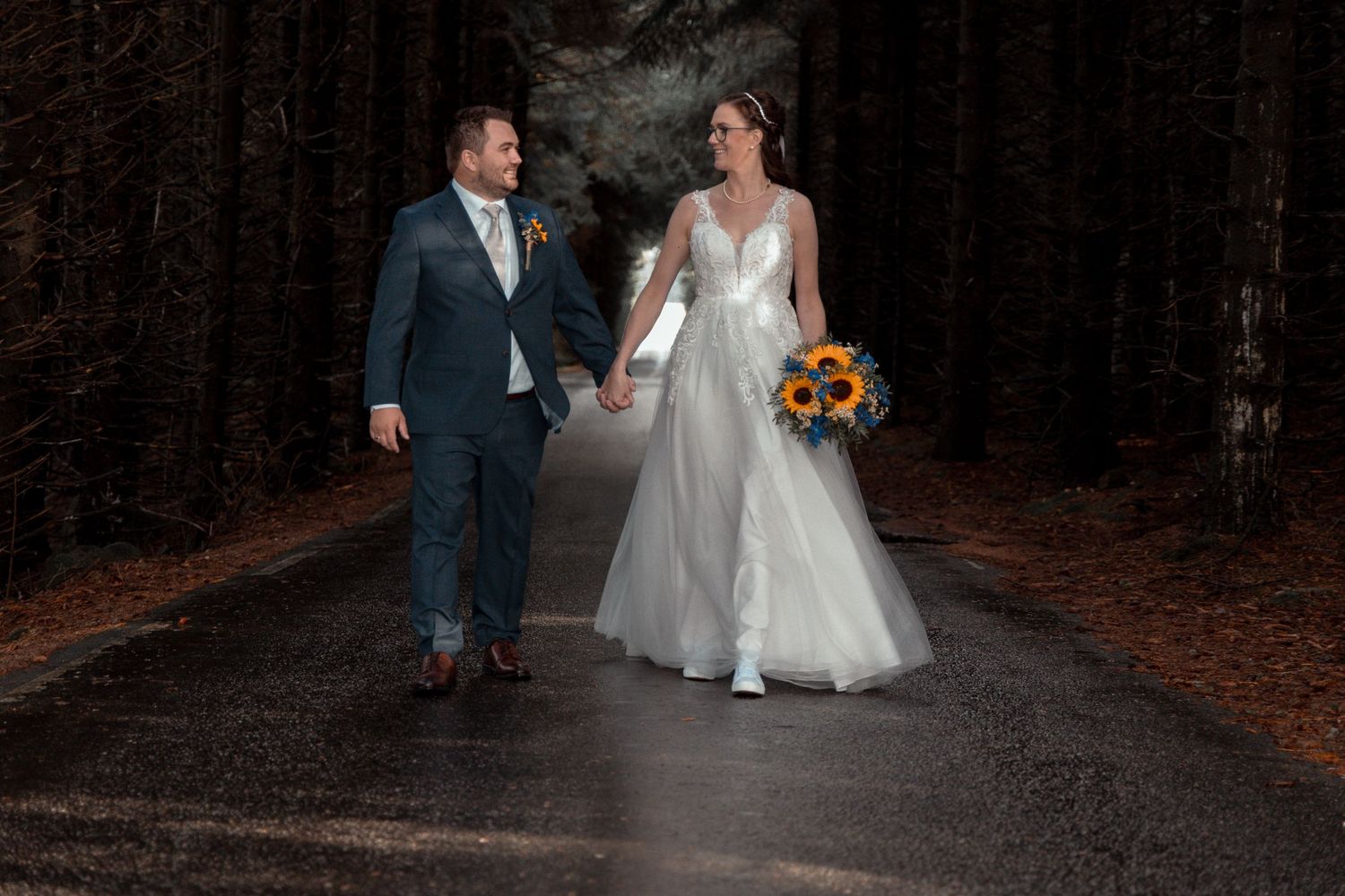 Bride and groom walking hand in hand on forest road with sunflower bouquet.