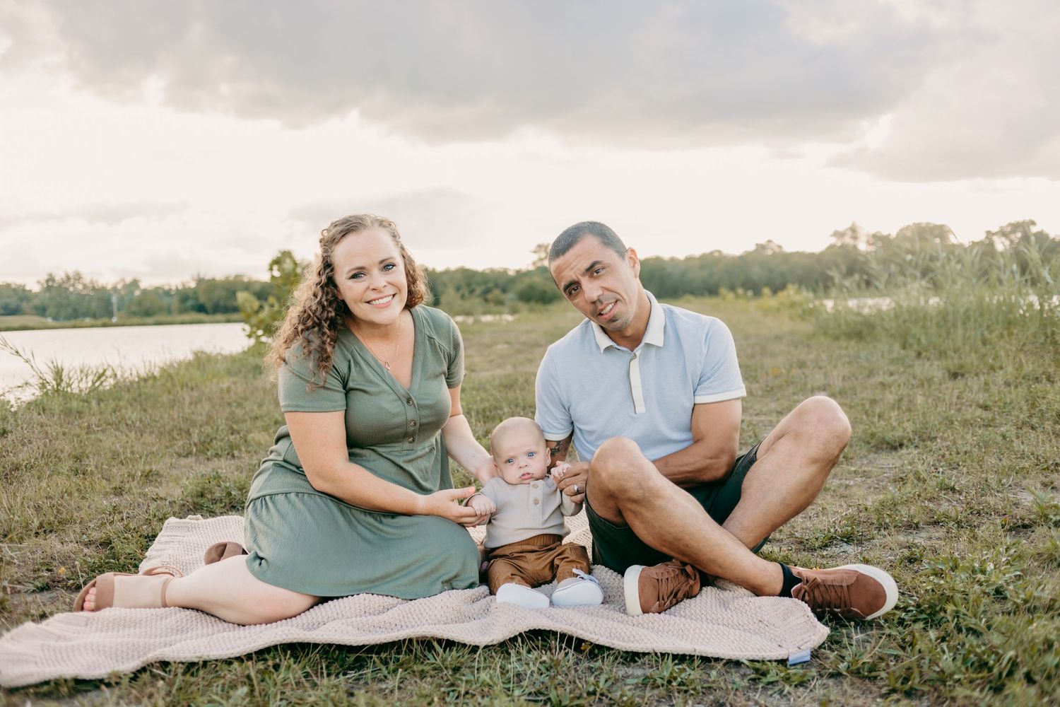 Family sitting together on blanket by lake during sunset photo session.