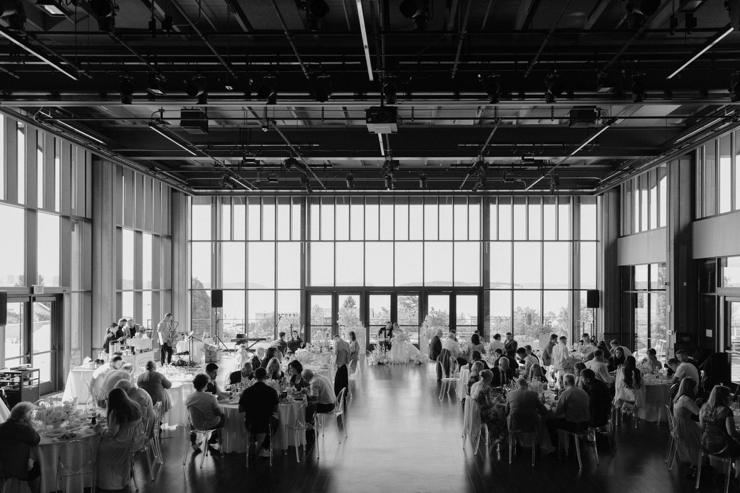 Black and white photo of a modern reception venue with large windows and guests seated at round tables.