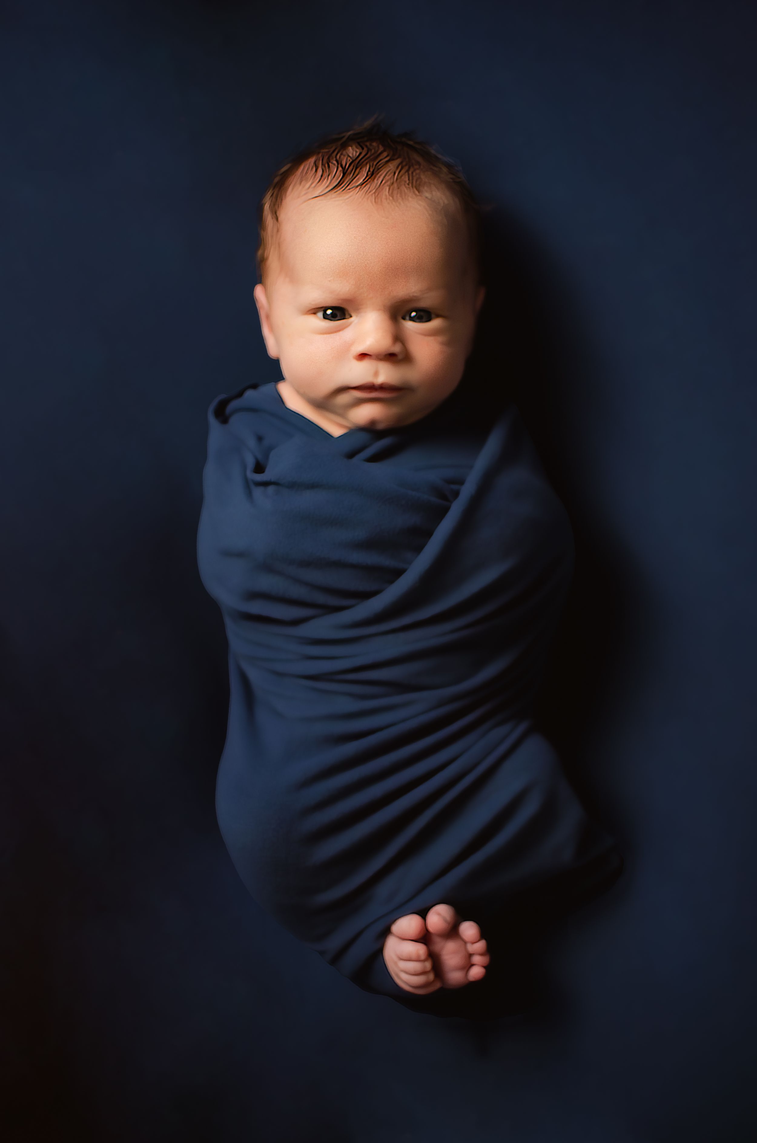 A newborn baby wrapped in navy blue fabric against a dark background.