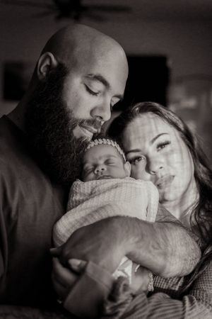 A tender black and white portrait of new parents cradling their sleeping newborn wrapped in a blanket.