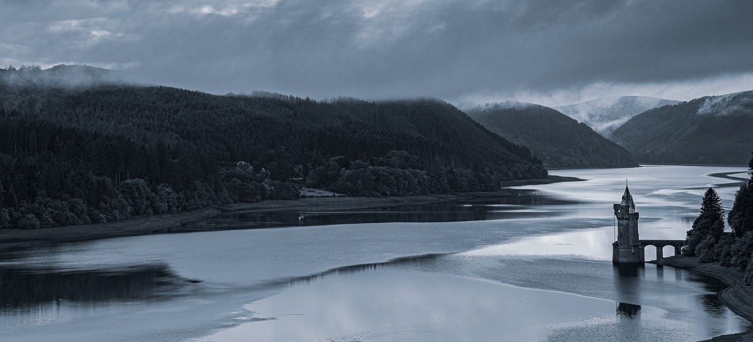 Misty mountain lake landscape with dark cloudy skies reflecting on calm waters.