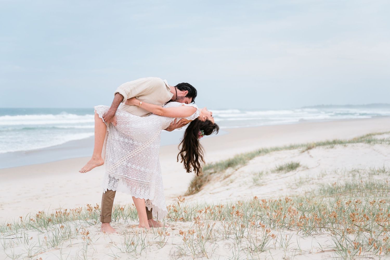A couple in white clothing embraces playfully on a sandy beach with ocean waves in the background.