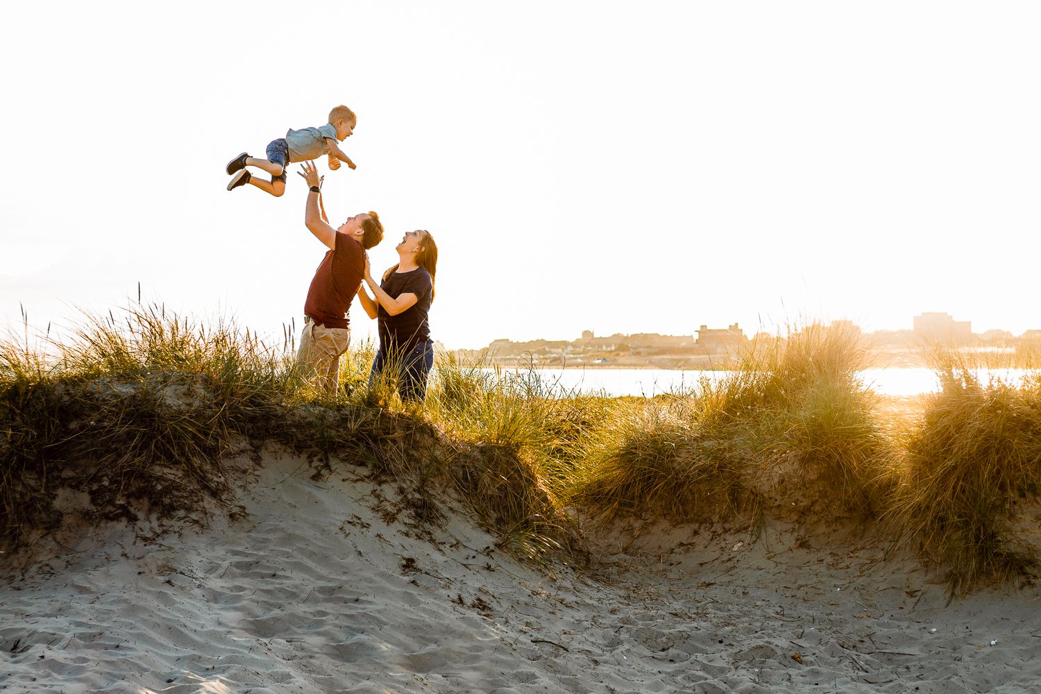 On a beach on Hayling Island a Father throws his three year old up in the air as the Mother looks on and laughs.