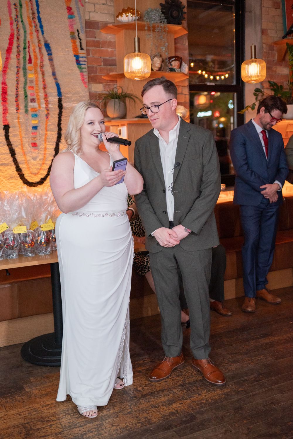 Couple in formal attire stands near a decorated bar with pendant lights and colorful wall decorations.