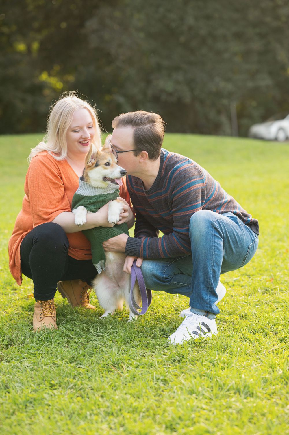 A couple crouches on grass while playing with their small dog on a sunny afternoon.