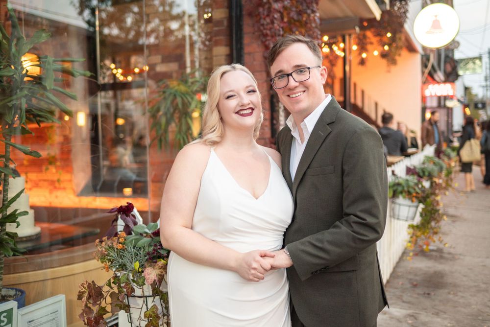 Elegant couple poses outside storefront with festive holiday decorations and lighting.