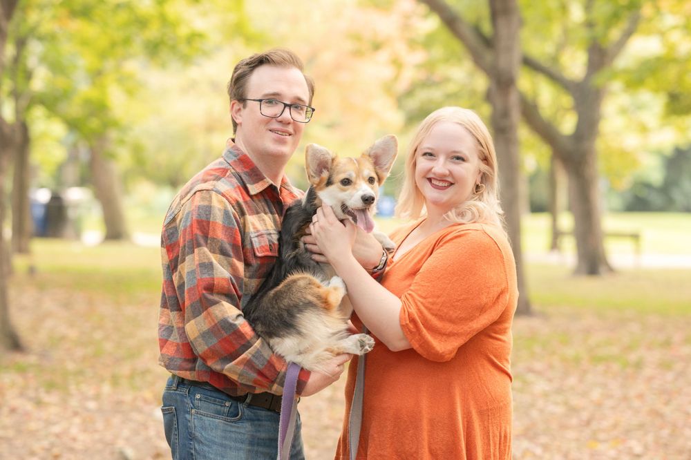 A couple poses with their Corgi dog amid autumn trees in a park setting.