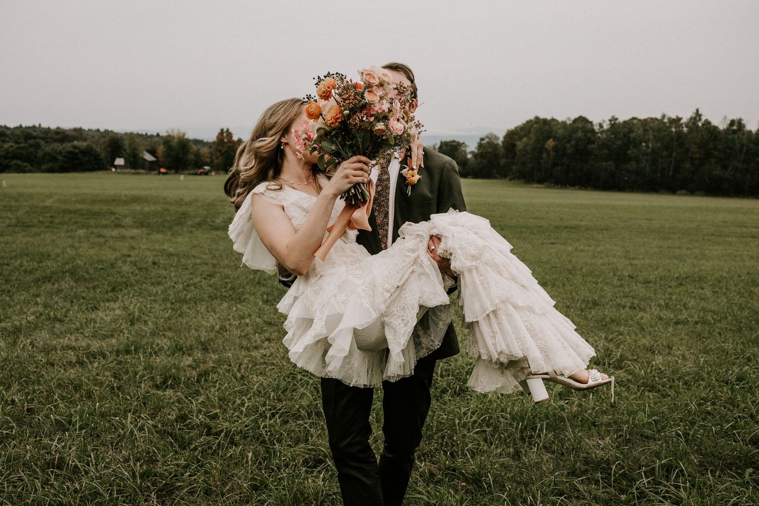 Groom carrying bride in white ruffled dress through green field, bride holding colorful flower bouquet. Newbury VT. Miranda Photo & Co