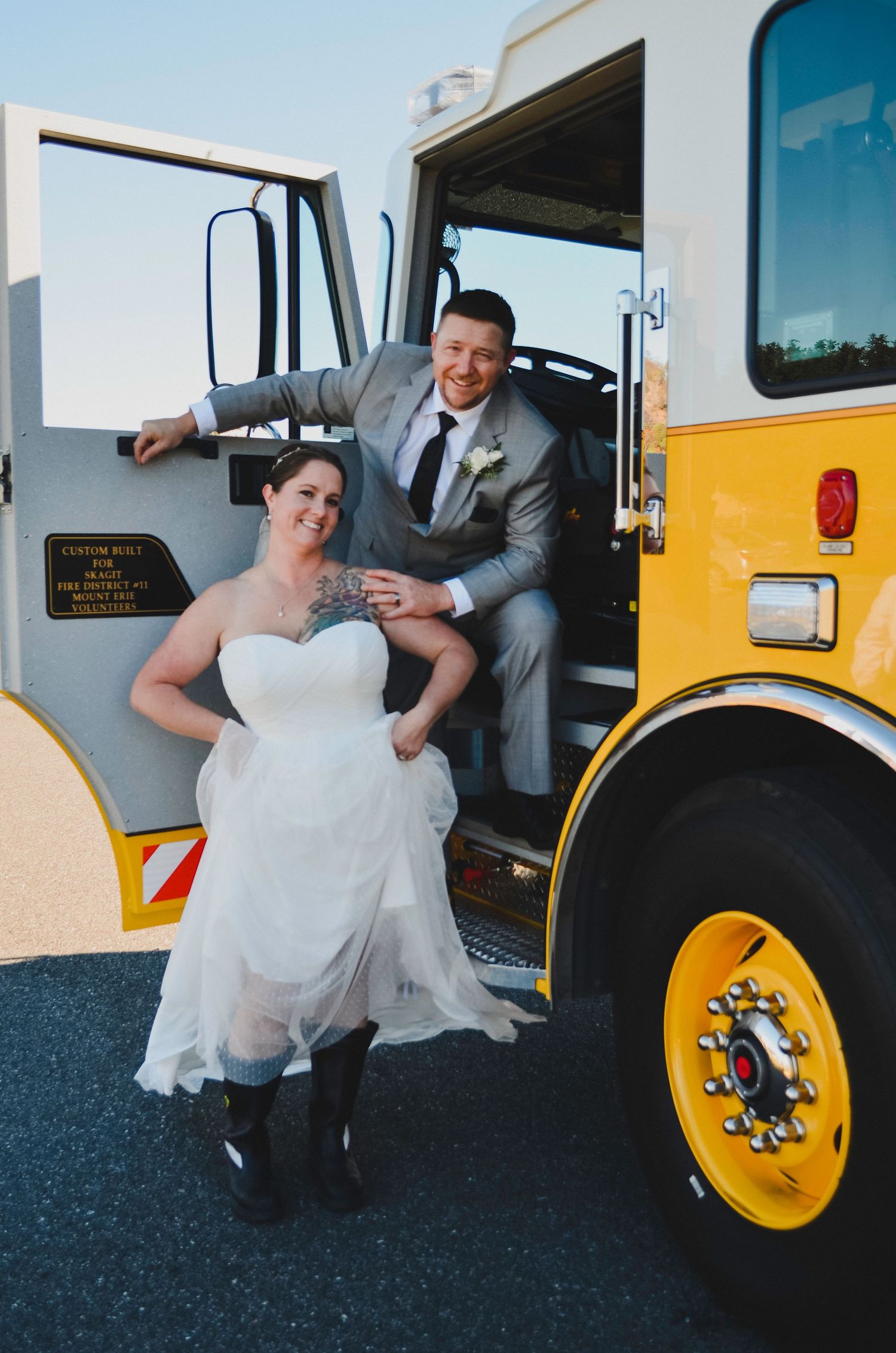 A bride in a white dress posing with someone in a grey suit by a yellow school bus on their wedding day.