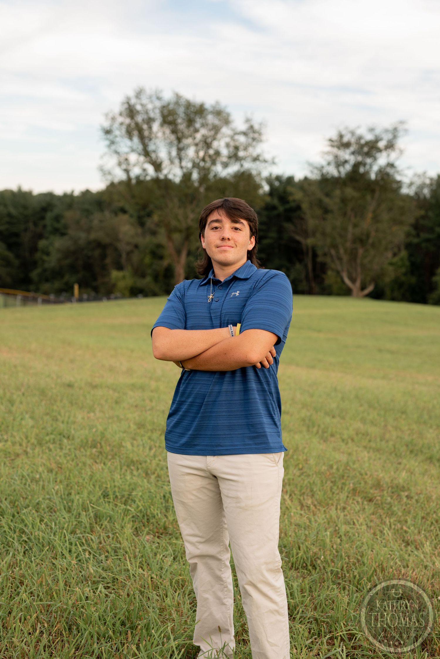 A person stands confidently with crossed arms in a blue polo and khakis on a grassy hillside.