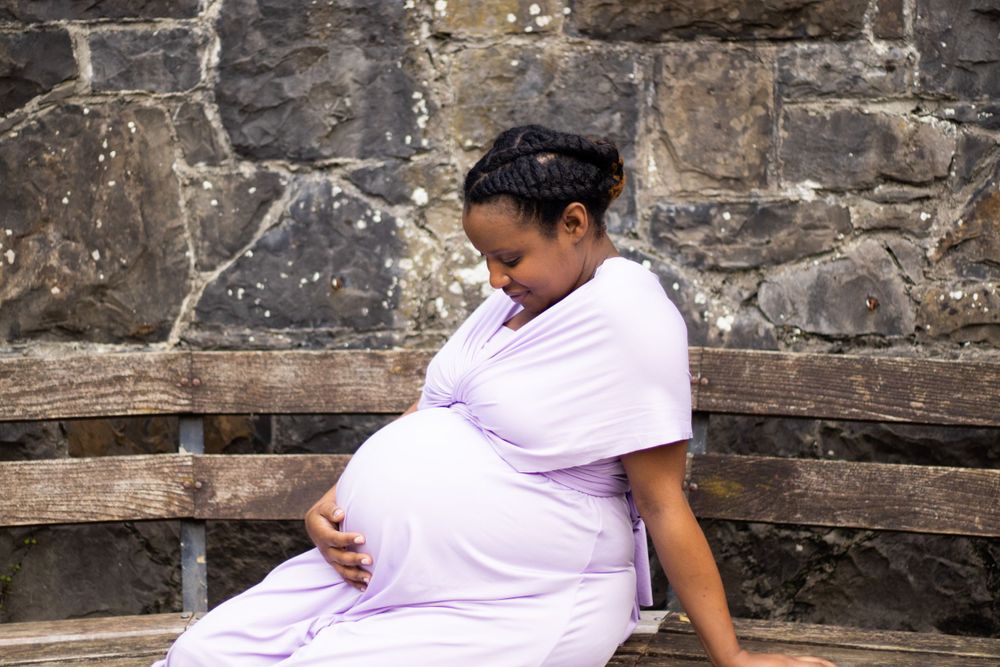 Expectant parent in lavender dress sitting on stone bench.